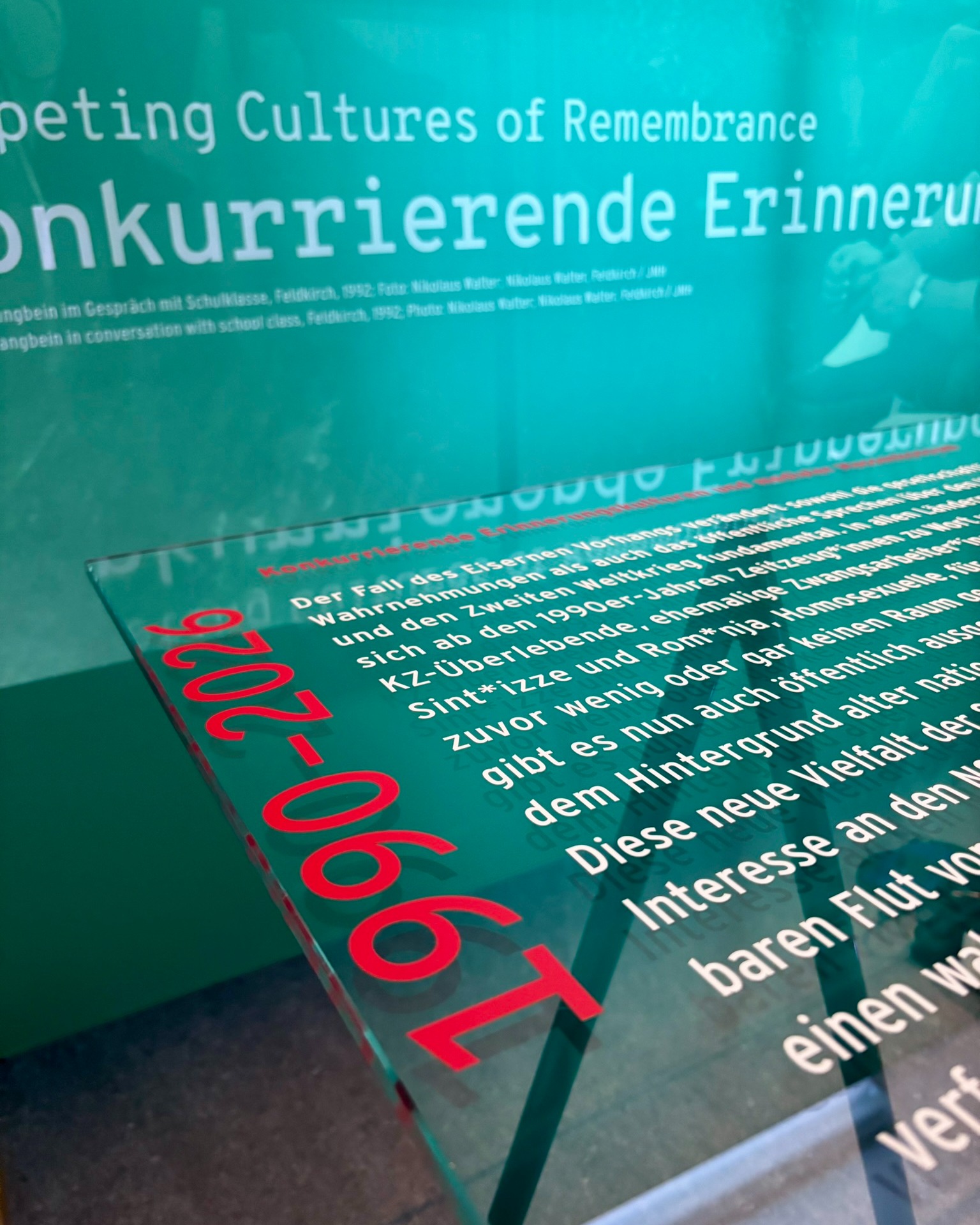 Ein Museumsinformationstafel mit dem Titel 'Competing Cultures of Remembrance' und 'Konkurrierende Erinnerungen' im Hintergrund. Im Vordergrund befindet sich eine schräg angebrachte Glasplatte mit der großen roten Jahreszahl '1990-2026'. Der weiße Text darauf erläutert den Fall des Eisernen Vorhangs und die gestiegene öffentliche Wahrnehmung von KZ-Überlebenden, Sinti und Roma sowie Homosexuellen. (KI-Alternativtext)