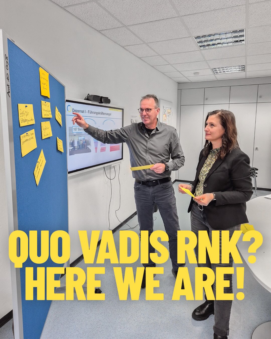 Dieser Alternativtext wurde KI-generiert: Ein Raum mit einer blauen Pinwand, auf der Post-its hängen. Zwei Personen stehen vor einem Bildschirm und diskutieren, wobei eine Person auf den Bildschirm zeigt.