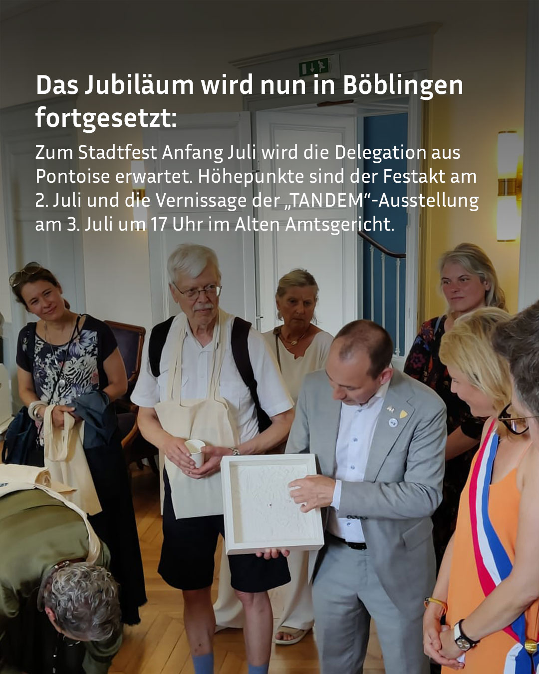 Eine Gruppe von Personen steht in einem Raum, wobei eine Person in einem grauen Anzug ein weißes, gerahmtes Objekt hält und einer anderen Person mit weißem Haar zeigt. Der eingeblendete Text lautet: „Das Jubiläum wird nun in Böblingen fortgesetzt: Zum Stadtfest Anfang Juli wird die Delegation aus Pontoise erwartet. Höhepunkte sind der Festakt am 2. Juli und die Vernissage der „TANDEM“-Ausstellung am 3. Juli um 17 Uhr im Alten Amtsgericht.“ (KI-Alternativtext)