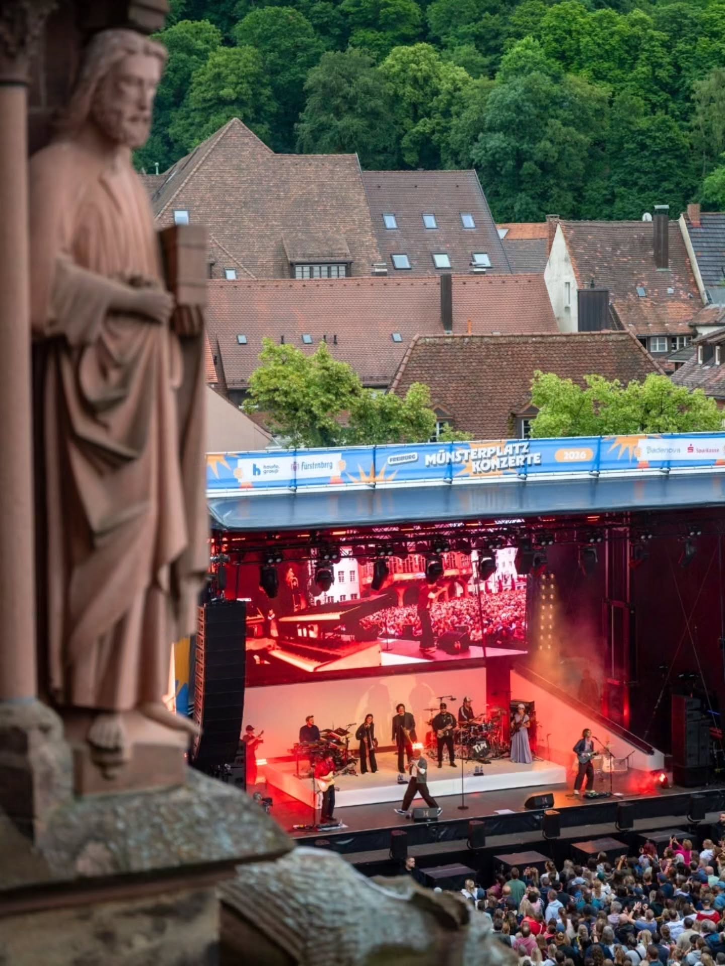 Eine Statue im Vordergrund blickt auf eine beleuchtete Bühne mit Musikern und einem großen Publikum; im Hintergrund sind Dächer und Bäume zu sehen, über der Bühne steht „Münsterplatz Konzerte 2024“. (KI-Alternativtext)