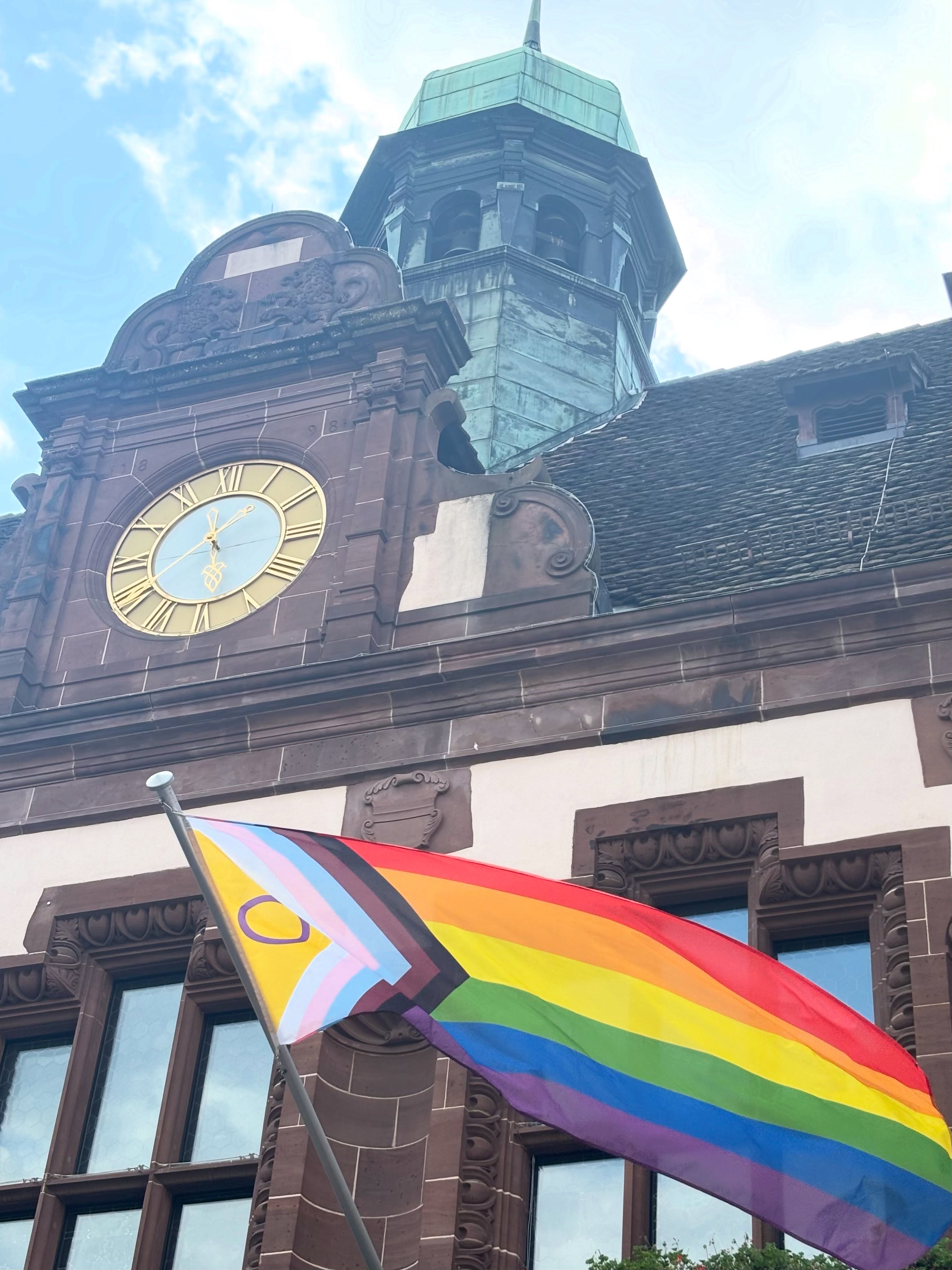 Ein historisches Gebäude mit Turm, grünem Kupferdach und großer Uhr mit römischen Ziffern. Im Vordergrund weht eine Progress Pride Flagge mit Regenbogenstreifen und einem Pfeil in Schwarz, Braun, Hellblau, Rosa und Weiß. (KI-Alternativtext)