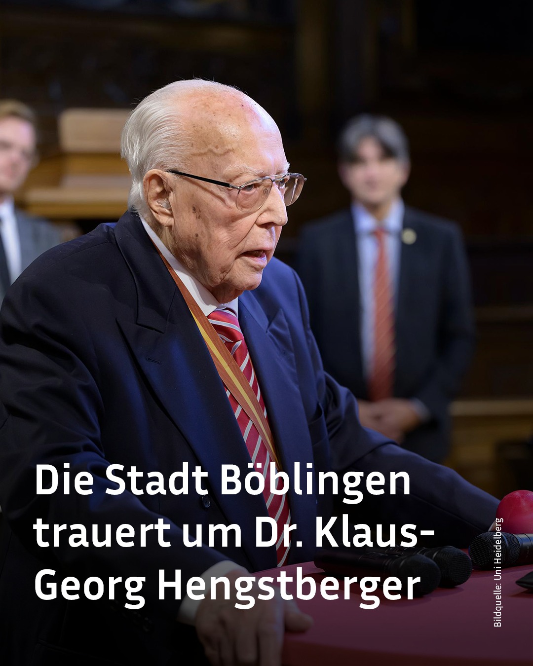 Ein älterer Mensch mit Brille und dunklem Anzug spricht an einem Rednerpult; im Hintergrund stehen zwei weitere Personen. Überlagert ist der Text: „Die Stadt Böblingen trauert um Dr. Klaus-Georg Hengstberger“. (KI-Alternativtext)