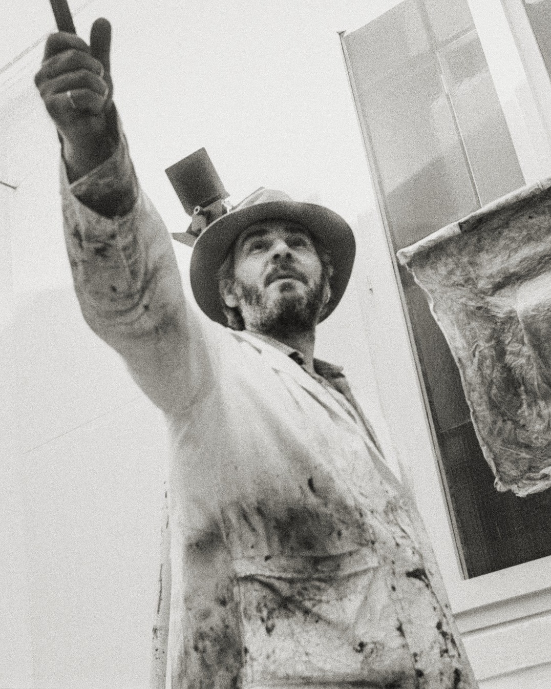 Dieser Alternativtext wurde KI-generiert: Schwarz-weißes Foto einer Person mit Hut und hochgehobener Hand. Im Hintergrund ist eine Fassade zu sehen.