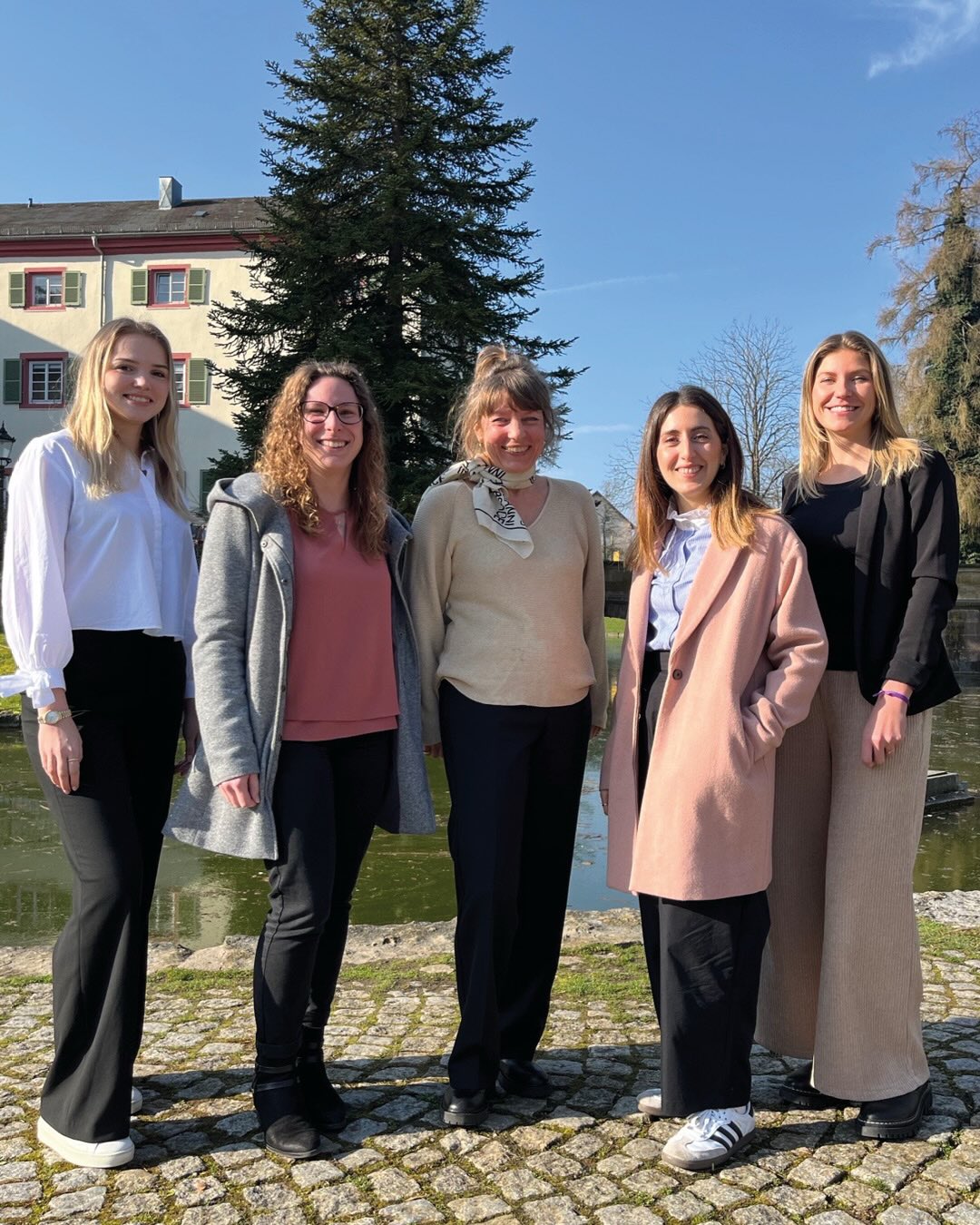 Dieser Alternativtext wurde KI-generiert: Eine Gruppe von Personen steht vor einem Gebäude und einem Teich. Im Hintergrund befindet sich ein großer Baum und ein klarer Himmel.