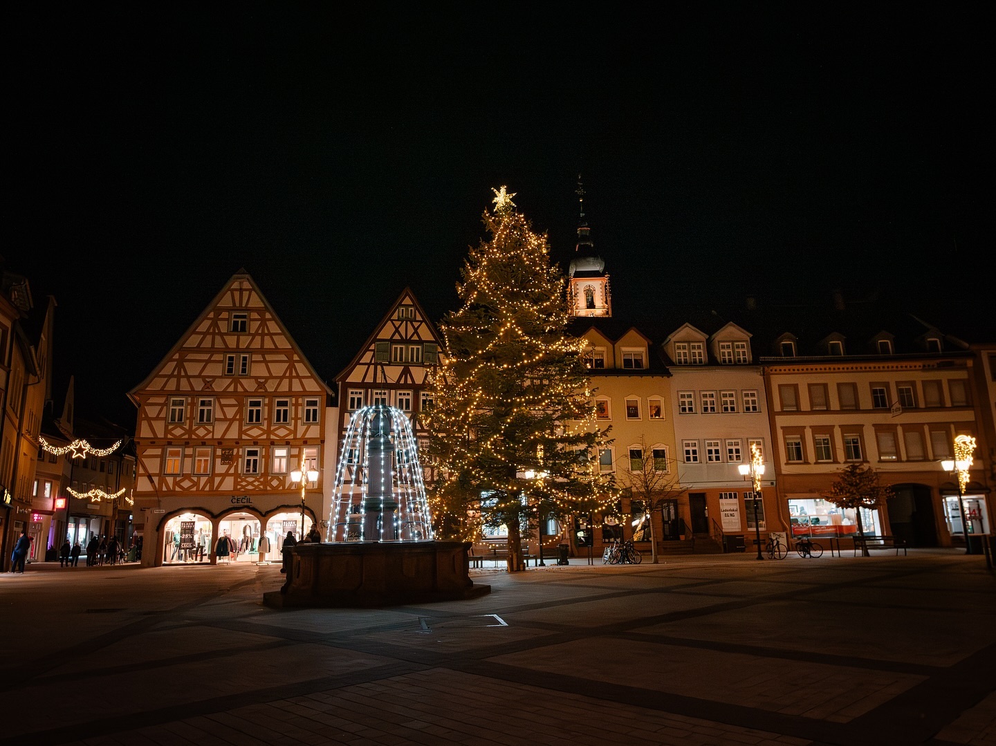 Ein beleuchteter Weihnachtsbaum steht auf einem Platz mit Fachwerkhäusern. Die Gebäude und der Brunnen sind ebenfalls weihnachtlich dekoriert.