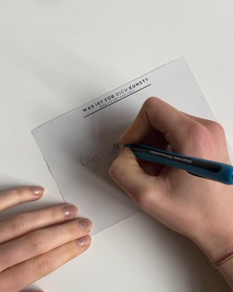 Dieser Alternativtext wurde KI-generiert: Ein Blatt Papier liegt auf einer Oberfläche und wird mit einem blauen Stift beschrieben. Auf dem Papier steht ein Text in deutscher und englischer Sprache.