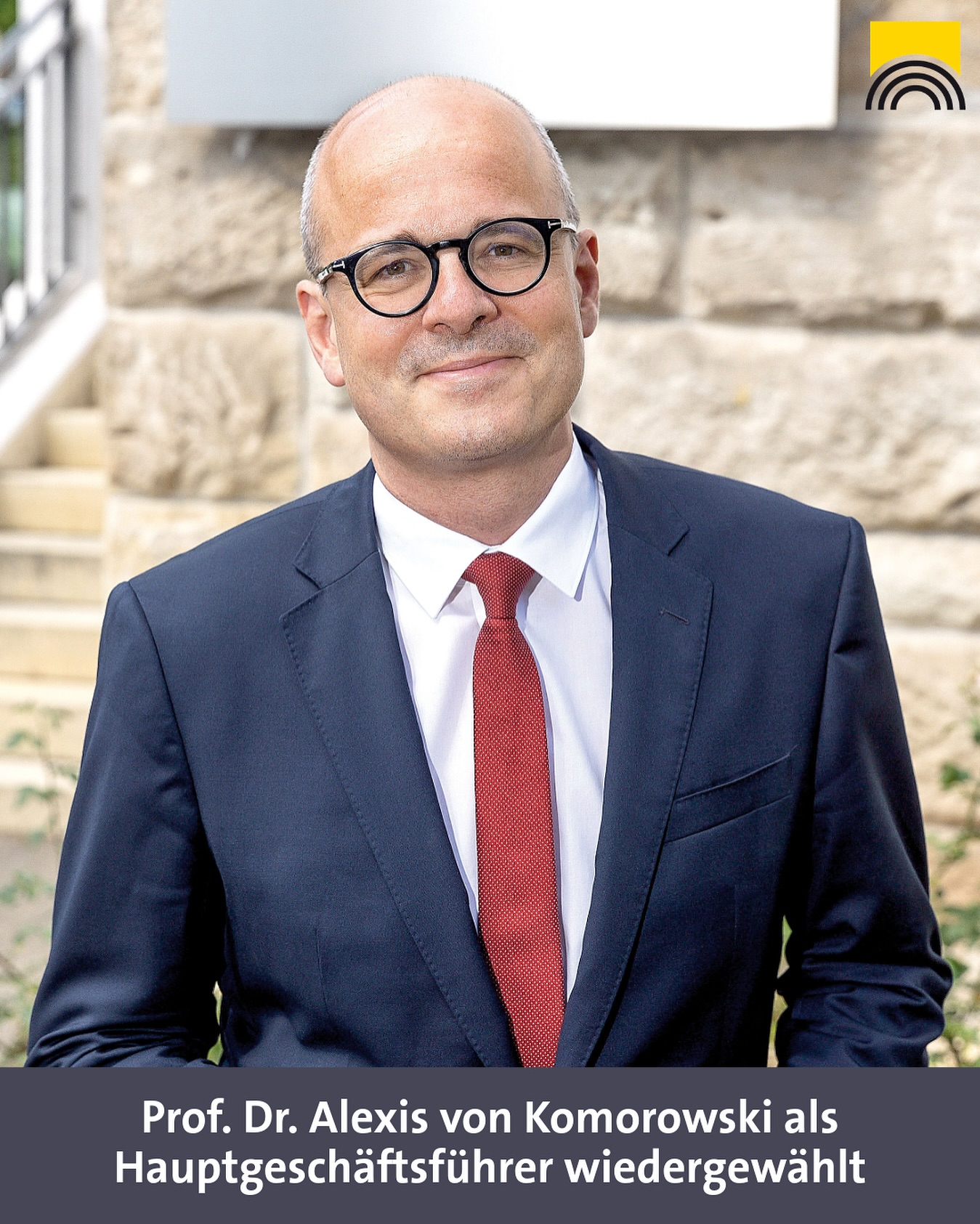 Ein Mensch mit Brille, Anzug und Krawatte posiert vor einer Wand. Im oberen Bereich ist ein Logo zu sehen.