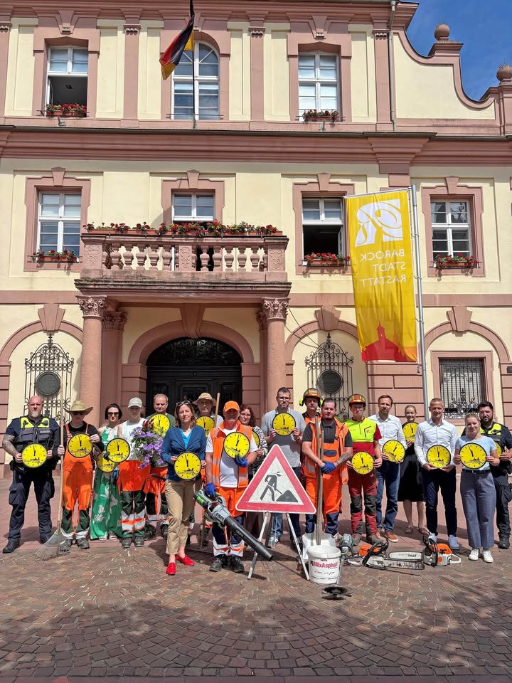 Gruppe von Menschen vor einem historischen Gebäude, die gelbe Schilder mit einer Uhr halten. Einige tragen Arbeitskleidung und stehen neben Baustellenschildern und Geräten. An der Fassade hängt eine deutsche Flagge und eine gelbe Fahne mit der Aufschrift „Barock Stadt Rastatt“. (KI-Alternativtext)