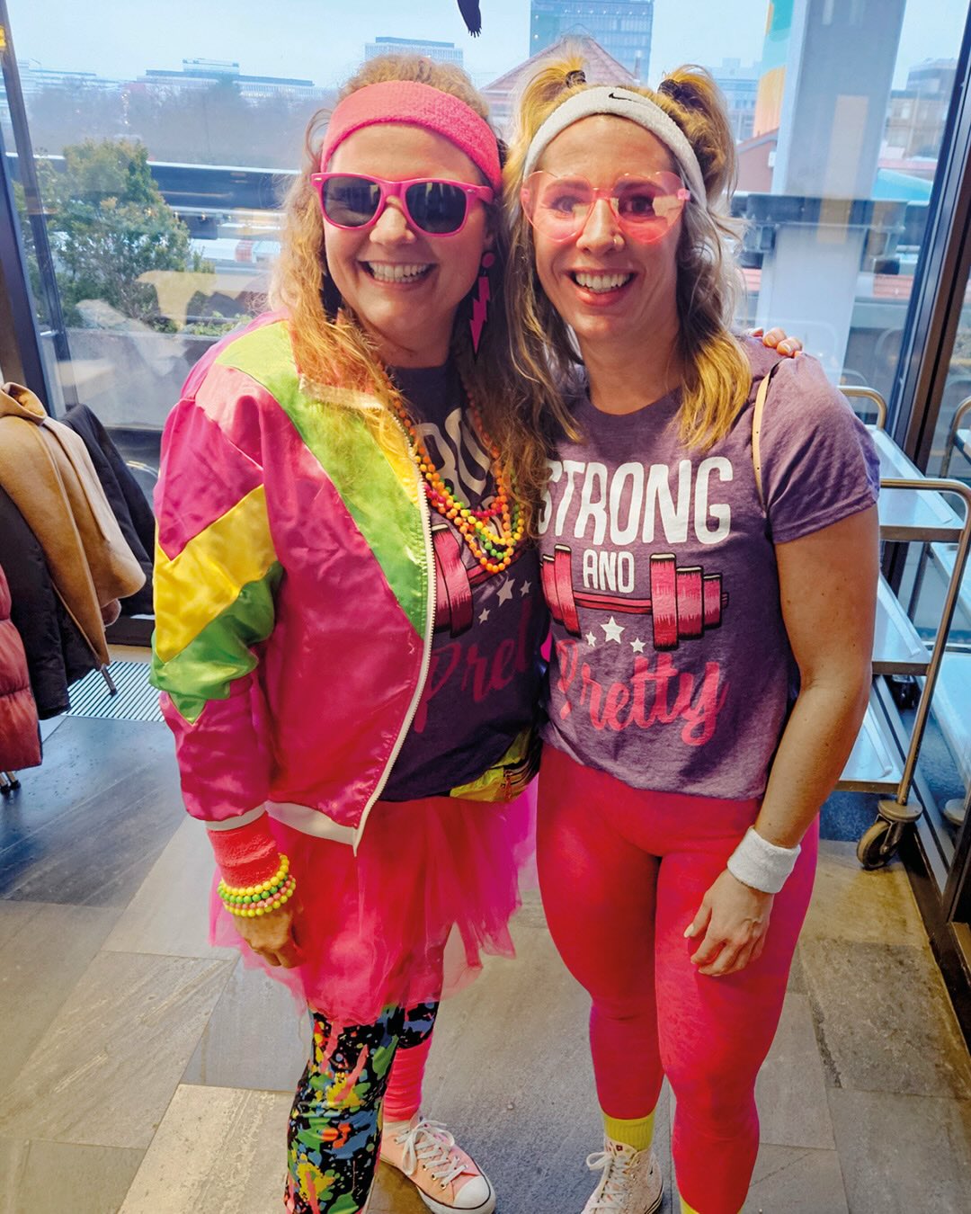 Dieser Alternativtext wurde KI-generiert: Zwei Personen lächeln in farbenfroher Kleidung und Accessoires. Im Hintergrund ist durch eine Glasscheibe eine Stadtlandschaft zu erkennen.