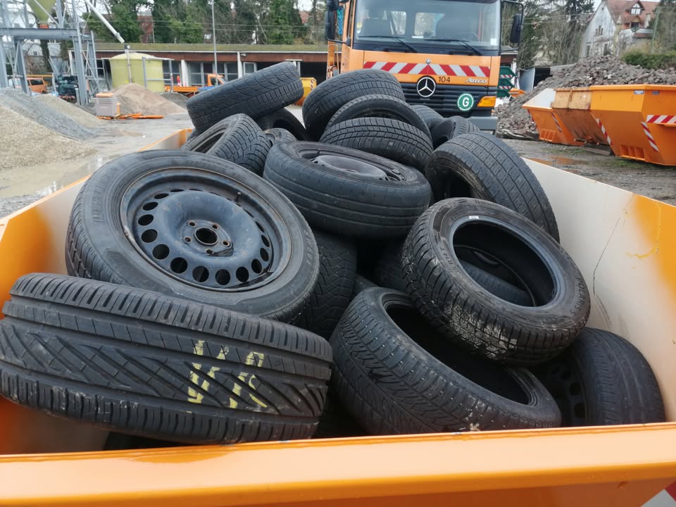 Dieser Alternativtext wurde KI-generiert: Eine Ansammlung gebrauchter Autoreifen liegt in einem orangefarbenen Behälter. Im Hintergrund sind Baumaschinen und eine Baustelle zu sehen.