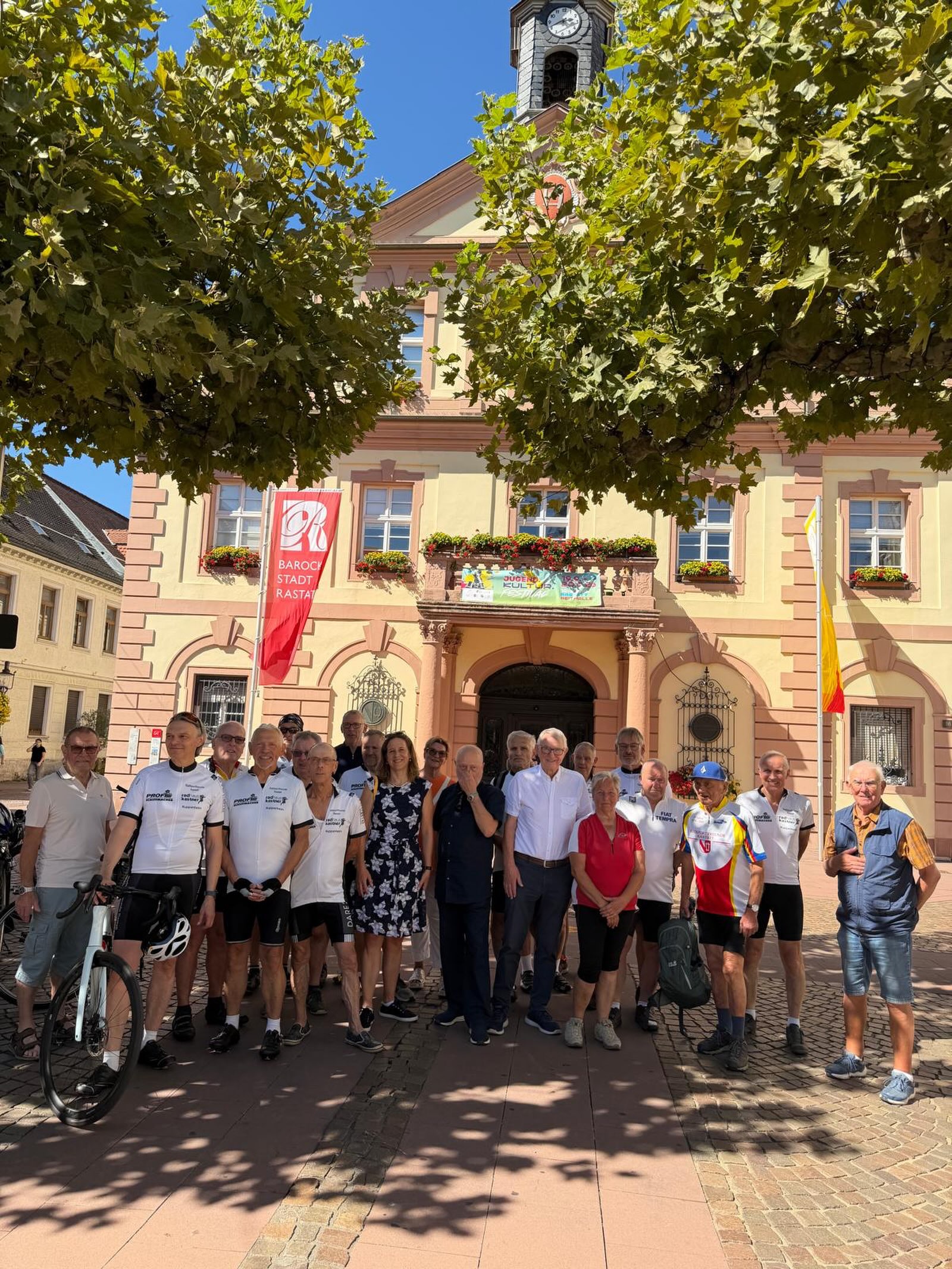 Eine große Gruppe von Menschen, viele davon in Radfahrerkleidung mit der Aufschrift "PROFIS", posiert vor einem gelben Gebäude mit Uhrturm. Links weht eine rote Flagge mit der Aufschrift "BAROCKSTADT RASTATT", während eine Person im Vordergrund ein Fahrrad hält. (KI-Alternativtext)
