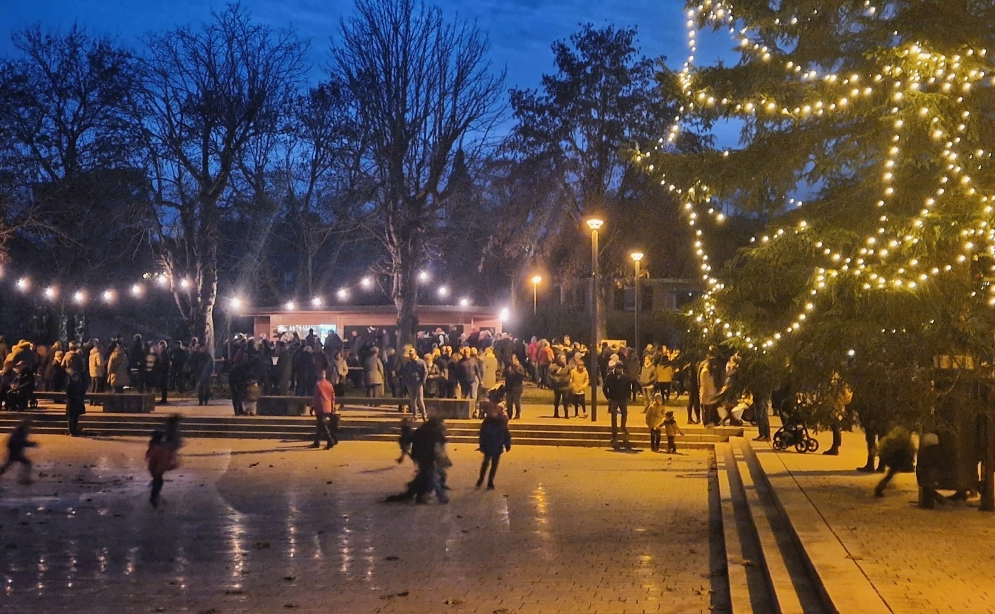 Ein beleuchteter Platz mit vielen Menschen, Bäumen und Lichtern. Es scheint ein festlicher Anlass zu sein.