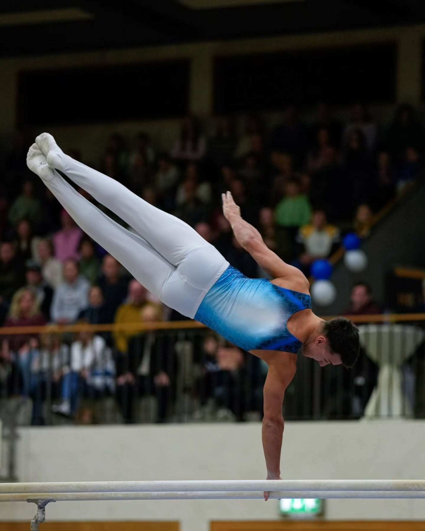 Dieser Alternativtext wurde KI-generiert: Eine Person führt eine akrobatische Übung auf einer Stange vor einem Publikum auf. Das Bild ist farbig und zeigt die Bewegung und Anstrengung der Performance.