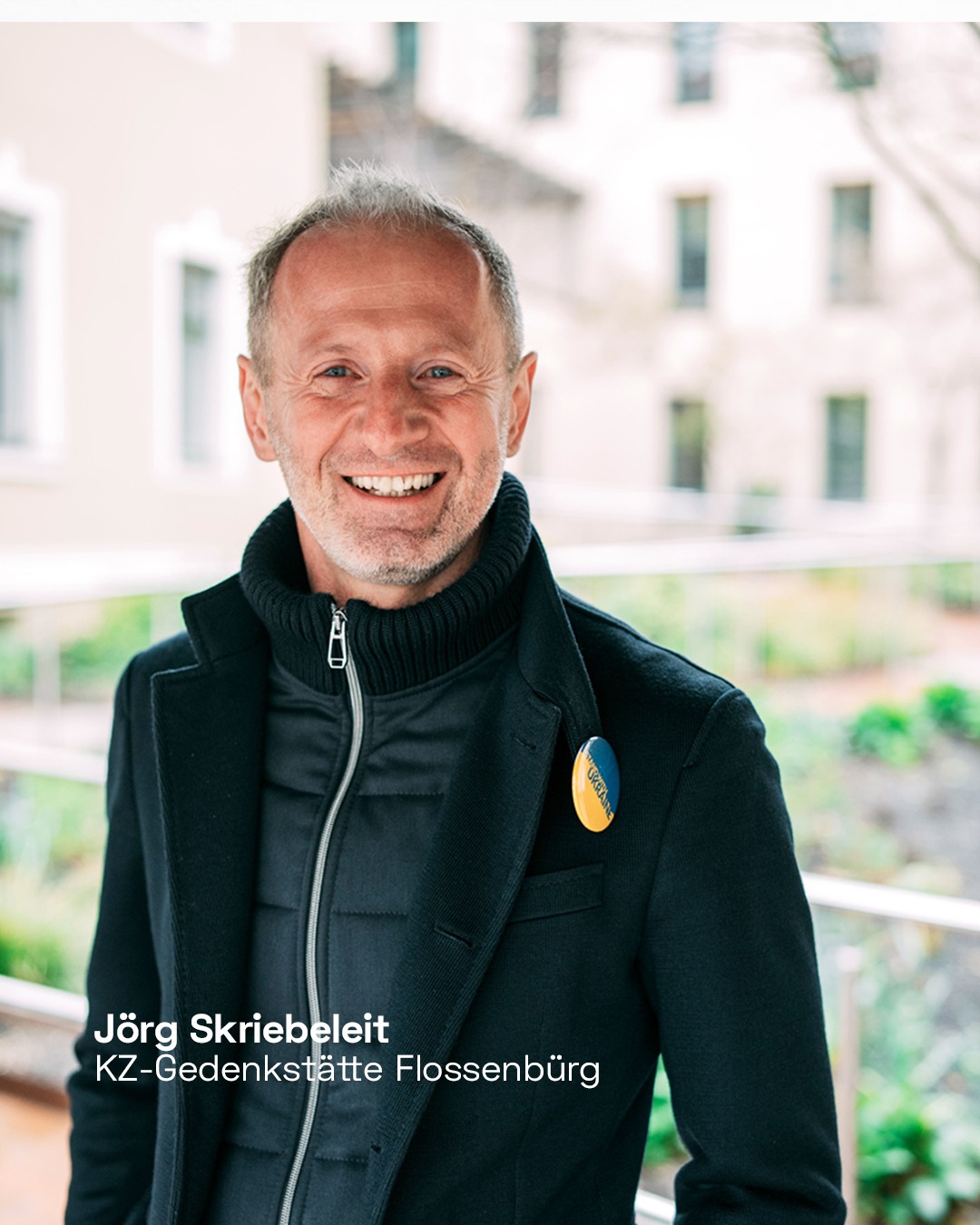 Eine lächelnde Person mit kurzem Haar trägt einen dunklen Mantel über einem Rollkragenpullover und ein rundes, gelb-grünes Abzeichen am Revers. Unten links steht der weiße Text „Jörg Skriebeleit KZ-Gedenkstätte Flossenbürg“. (KI-Alternativtext)