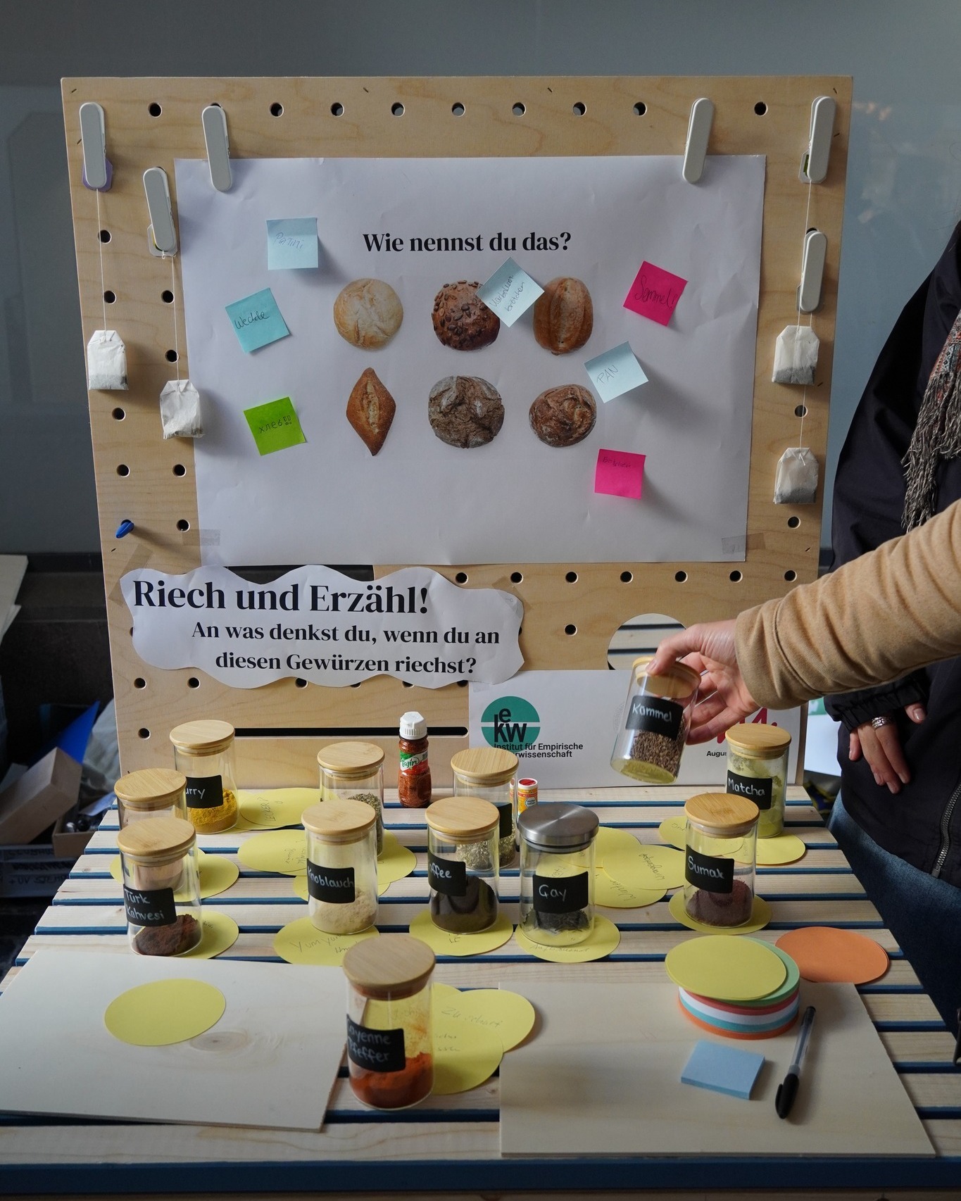 Dieser Alternativtext wurde KI-generiert: Eine Lochwand mit Post-its und verschiedenen Objekten, darunter Lebensmittel und kleine Flaschen. Darunter steht ein Tisch mit weiteren Flaschen und Tellern, vermutlich Teil einer Ausstellung zum Thema Gerüche und Gewürze.