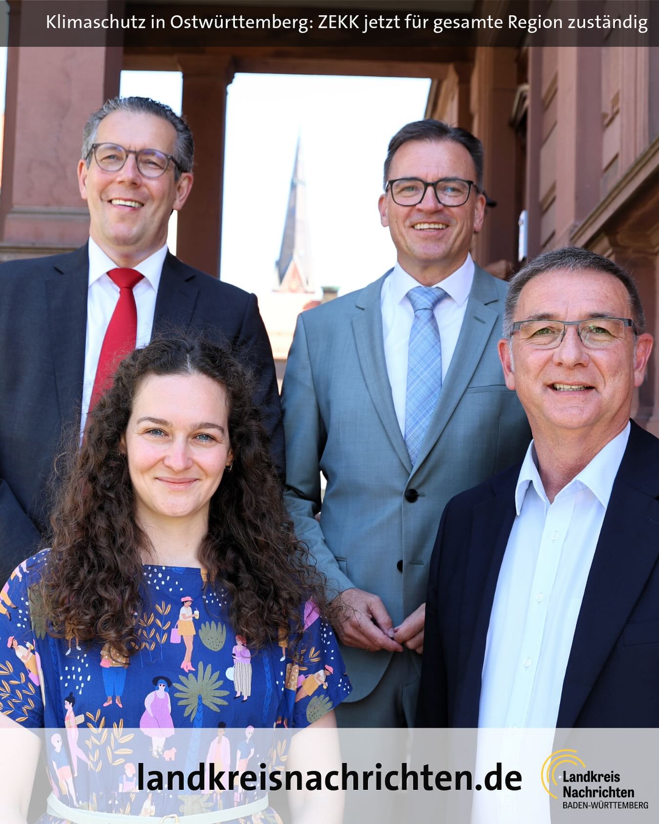 Ein Gruppenfoto von vier lächelnden Personen. Es ist eine Außenaufnahme vor einem Gebäude.