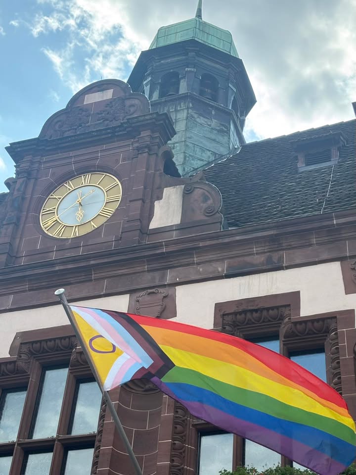 Eine Flagge mit Regenbogenstreifen sowie zusätzlichen Streifen in Schwarz, Braun, Hellblau, Pink und Weiß weht vor einem historischen Gebäude. Das Gebäude verfügt über einen Turm mit grünem Dach und eine große goldene Uhr mit römischen Ziffern. Der Himmel ist blau und teilweise bewölkt. (KI-Alternativtext)