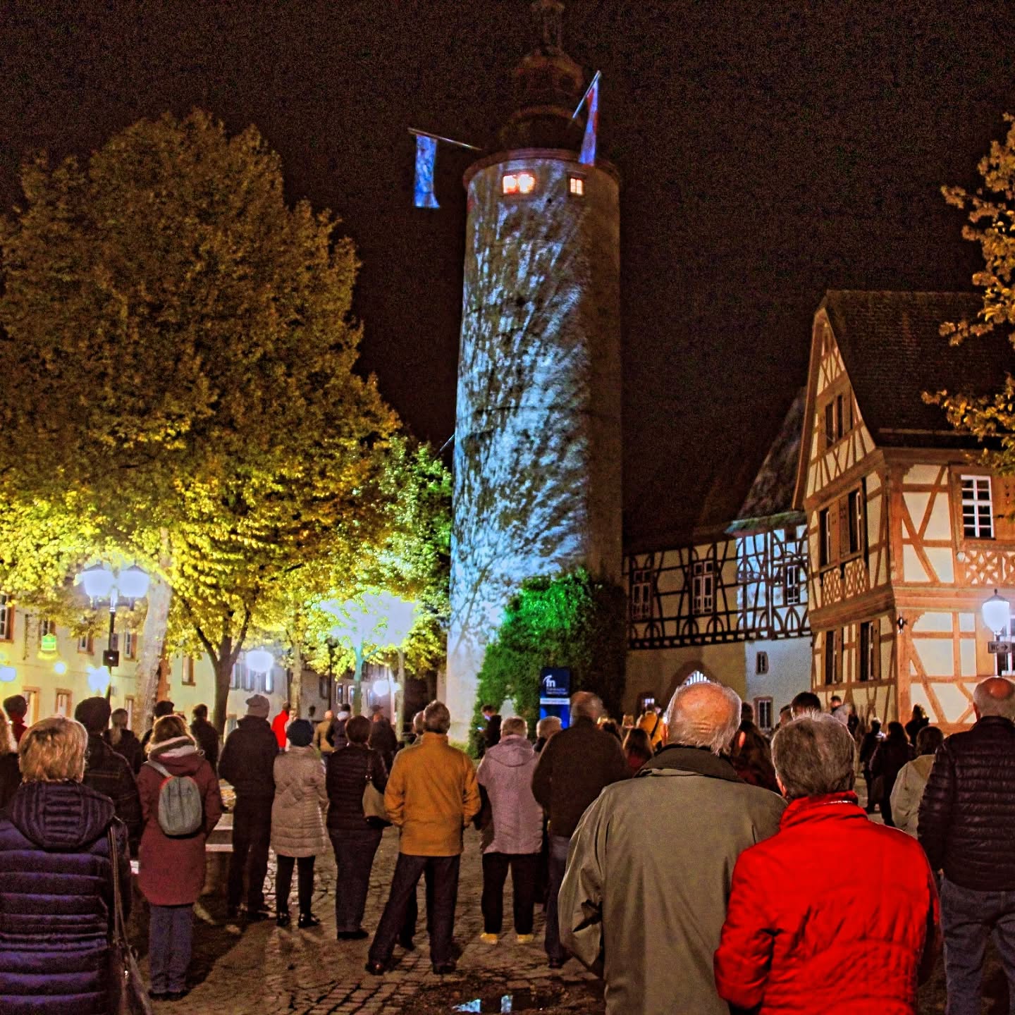 Dieser Alternativtext wurde KI-generiert: Nachts aufgenommenes Bild einer beleuchteten Turmstruktur, umgeben von Gebäuden und einer Menschenmenge. Das Bild ist farbig und zeigt eine stimmungsvolle Szene in einer Stadt.