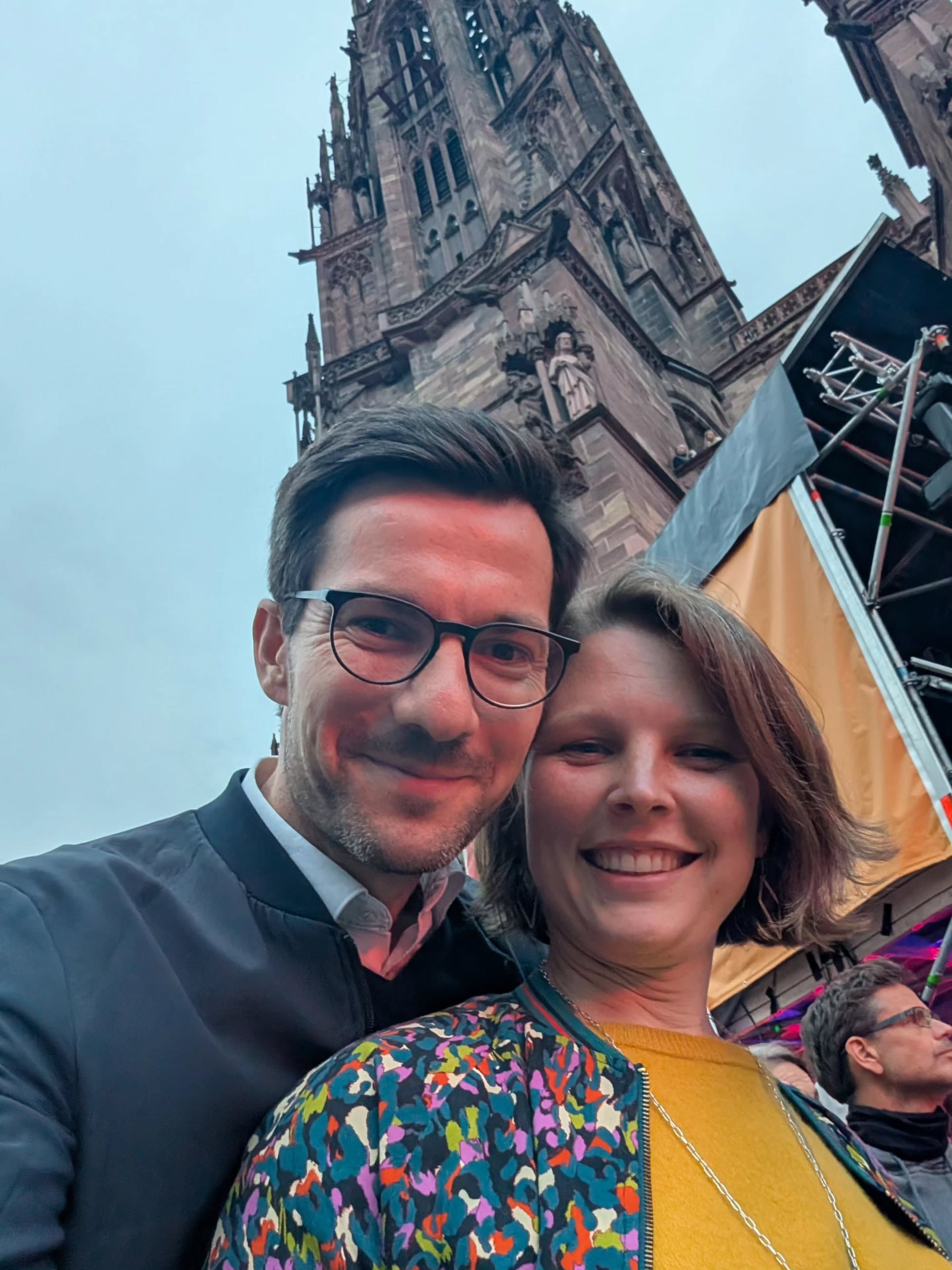 Zwei Personen lächeln in die Kamera, im Hintergrund eine große gotische Kirche mit Turm und ein Teil einer Bühne mit Gerüst. Eine Person trägt eine Brille und eine dunkle Jacke, die andere eine bunt gemusterte Jacke über einem gelben Oberteil. (KI-Alternativtext)