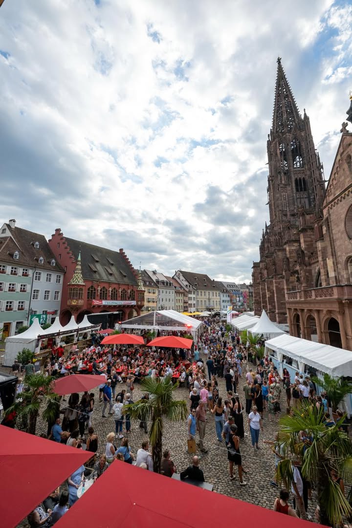 Ein überlaufener Marktplatz mit historischer Architektur, darunter ein hoher gotischer Kirchturm rechts und giebelständige Häuser links. Zahlreiche Menschen flanieren zwischen weißen Festzelten, roten Sonnenschirmen und Palmen, was auf ein Stadtfest hindeutet. (KI-Alternativtext)