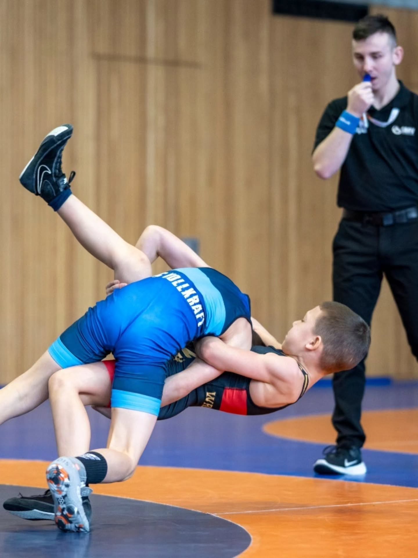 Dieser Alternativtext wurde KI-generiert: Zwei Personen ringen auf einer blauen Matte. Eine Person trägt blaue und die andere schwarze Sportkleidung.