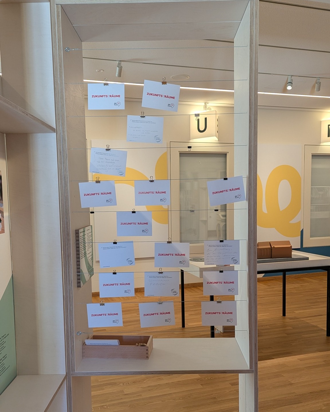 Dieser Alternativtext wurde KI-generiert: Ein Innenraum mit mehreren Informationen an einer durchsichtigen Trennwand aufgehängt. Sichtbare Dokumente sind mit Büroklammern befestigt.