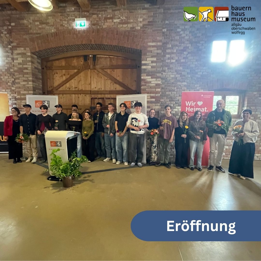 Dieser Alternativtext wurde KI-generiert: Eine Gruppe von Personen steht in einem Raum mit Backsteinwänden. Im Hintergrund befindet sich eine große, offene Tür und ein Banner mit der Aufschrift "Wir Heimat."