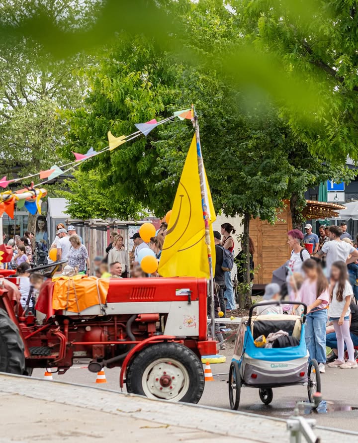 Dieser Alternativtext wurde KI-generiert: Eine verschwommene Szene zeigt eine Straßenparade mit bunten Flaggen und Luftballons. Im Vordergrund ist ein roter Traktor zu sehen, der von Menschen umgeben ist.