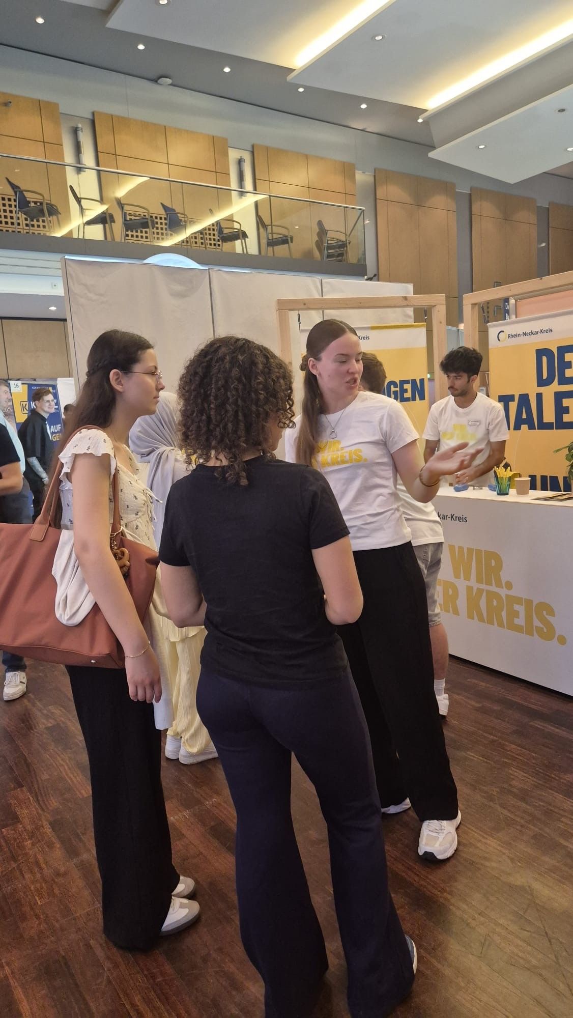 Mehrere Personen stehen in einem hellen Veranstaltungsraum vor einem Informationsstand und unterhalten sich. Eine Person im weißen T-Shirt mit gelber Schrift gestikuliert während des Gesprächs. Im Hintergrund sind Banner mit den Aufschriften „Rhein-Neckar-Kreis“, „DEIN TALENT“ und „WIR. ...R KREIS.“ zu sehen. (KI-Alternativtext)