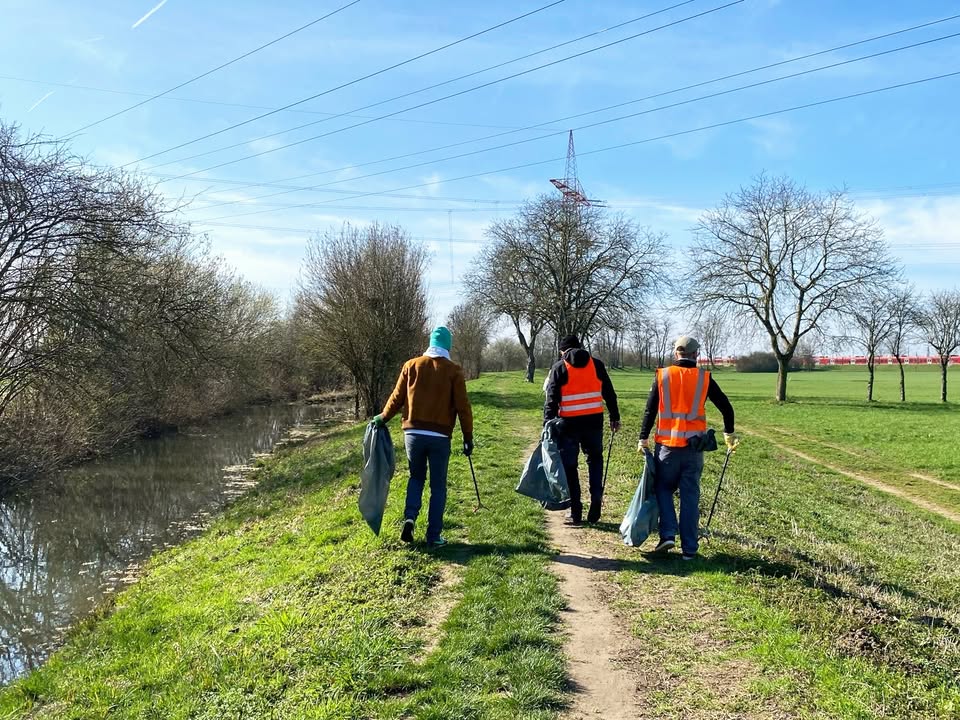 Dieser Alternativtext wurde KI-generiert: Mehrere Personen gehen auf einem Pfad entlang eines Wasserwegs, ausgestattet mit Westen und Tragetaschen. Im Hintergrund erstrecken sich Felder und Strommasten unter einem blauen Himmel.