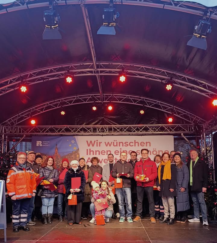 Dieser Alternativtext wurde KI-generiert: Eine Gruppe von Menschen steht vor einem Bühnenaufbau mit weihnachtlicher Dekoration. Ein Banner mit Text ist im Hintergrund zu sehen.