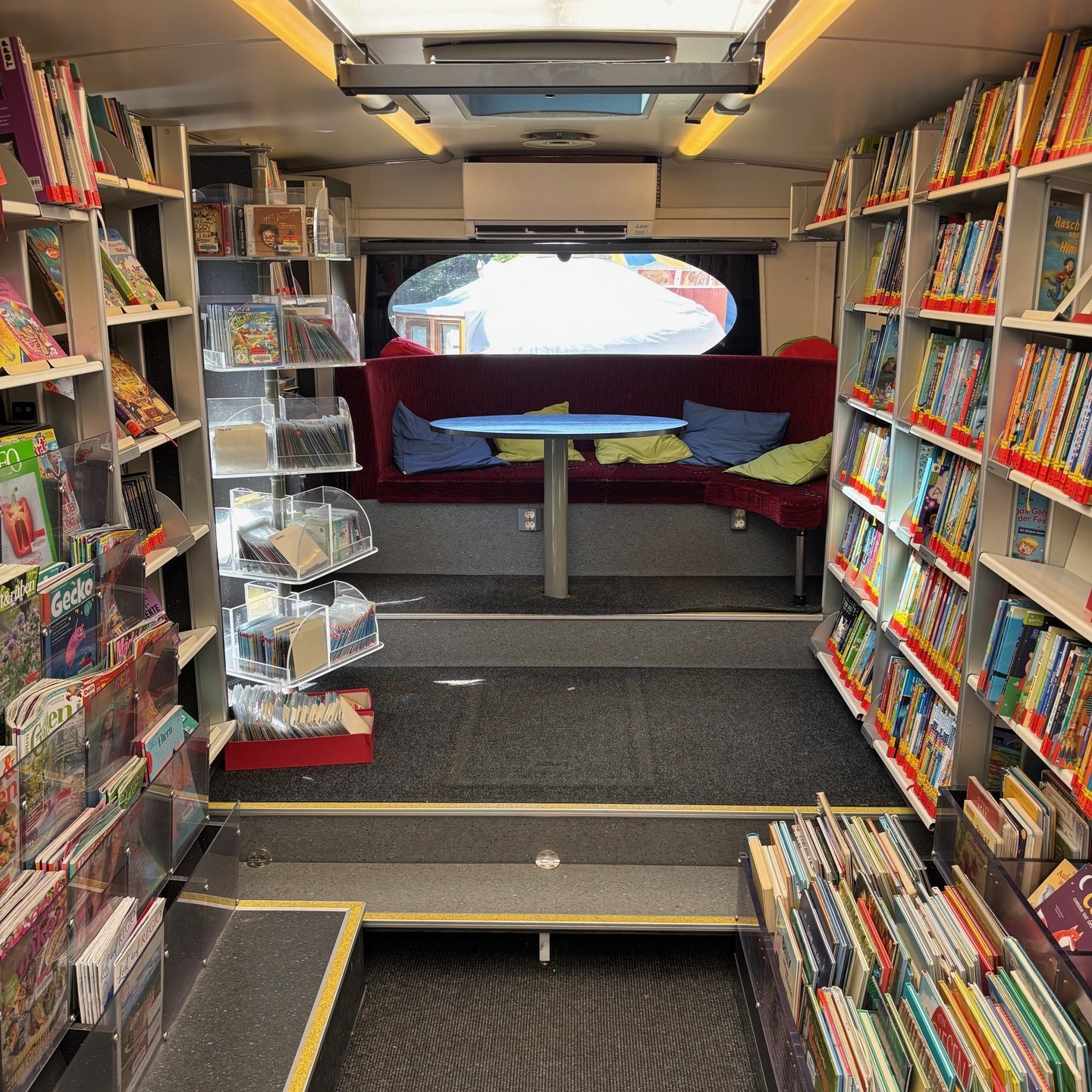 Das Bild zeigt das Innere eines Fahrzeugs. Es gibt Regale voller Bücher und einen Sitzbereich mit einem Tisch.
