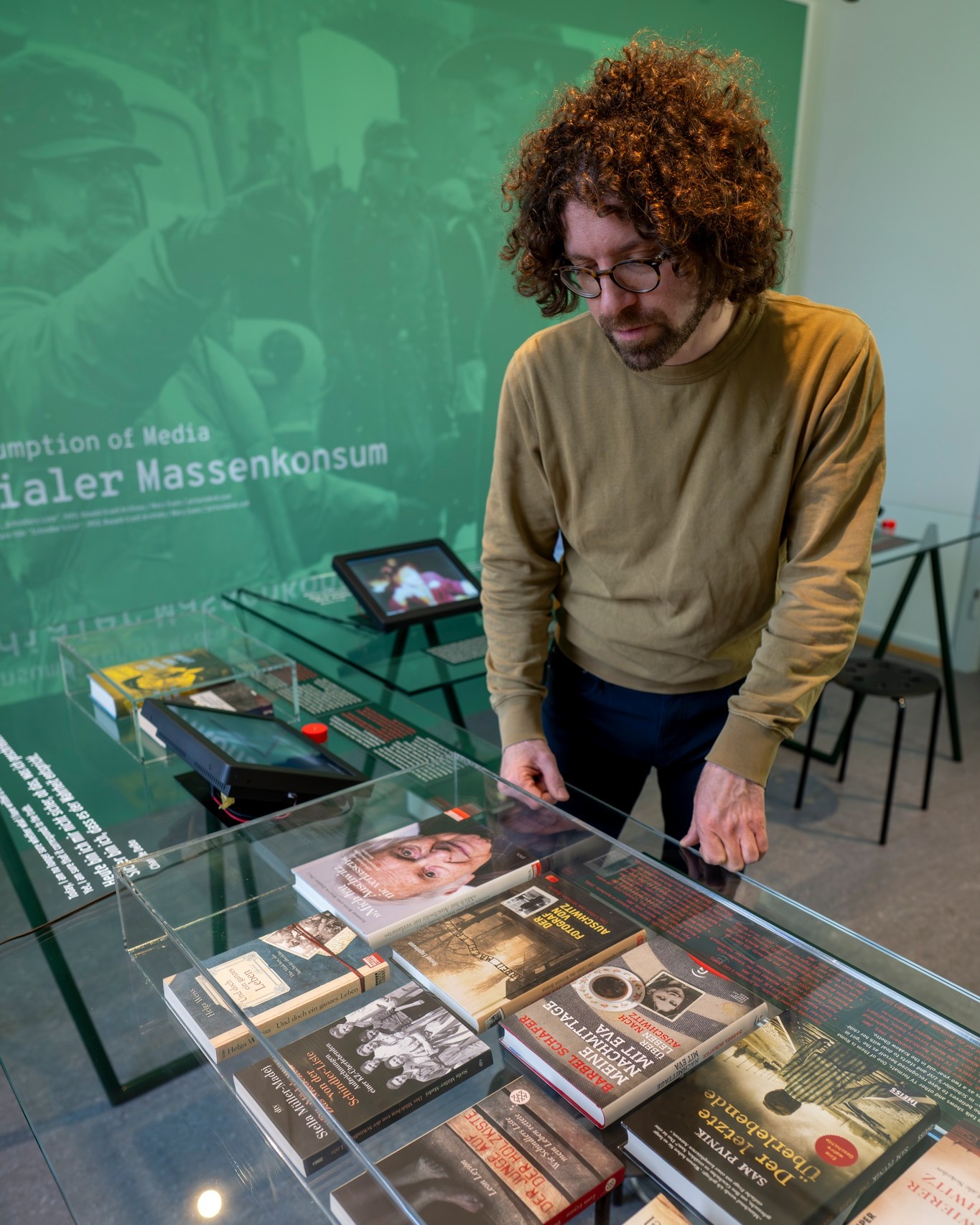 Person mit lockigem Haar und Brille betrachtet Bücher in einer Glasvitrine vor einer grünen Wand mit dem Text „Digitaler Massenkonsum“. Zu den sichtbaren Buchtiteln gehören „Auschwitz“, „Der Fotograf von Auschwitz“ und „Meine Nachmittage mit Eva“. (KI-Alternativtext)