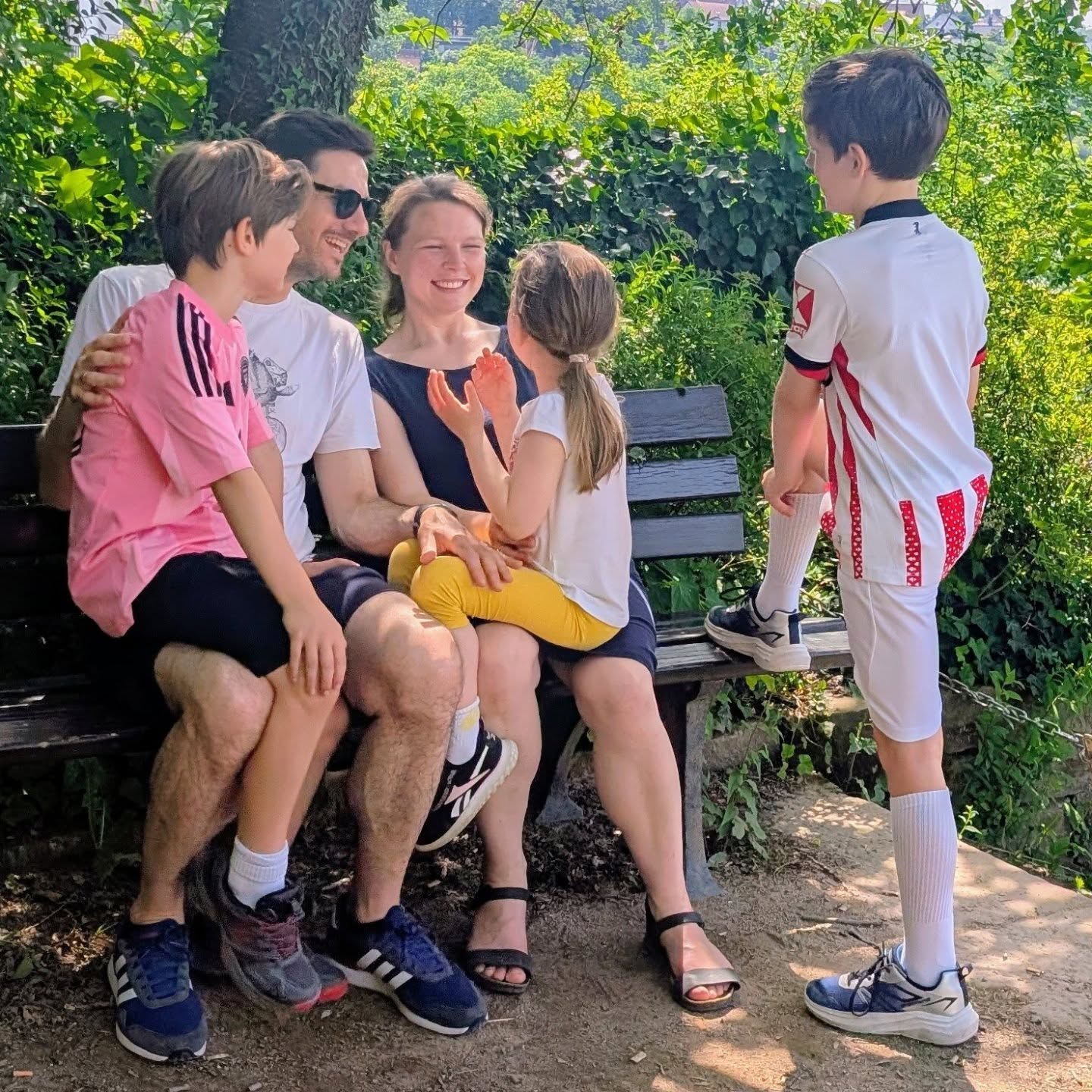 Eine Familie mit drei Kindern sitzt und steht lächelnd auf einer Parkbank vor grüner Vegetation. Ein Kind sitzt auf dem Schoß einer erwachsenen Person, ein anderes steht daneben und stützt sich auf die Bank. Alle tragen Freizeitkleidung, eines der Kinder trägt ein Fußballtrikot. (KI-Alternativtext)