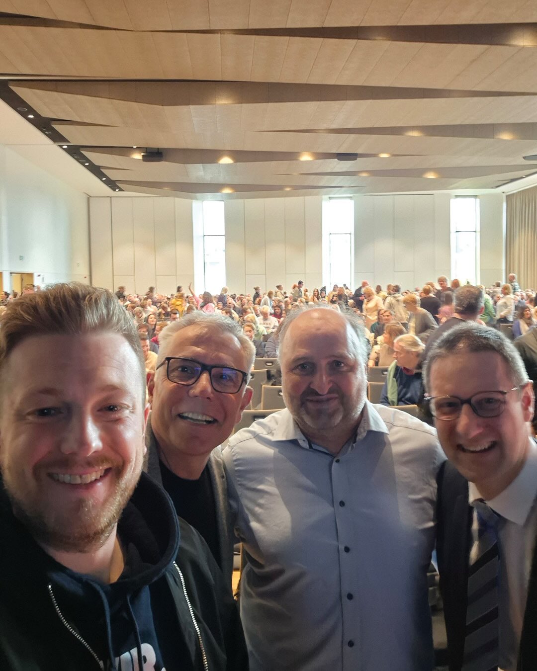 Dieser Alternativtext wurde KI-generiert: Ein farbiges Foto zeigt eine Menschenmenge in einem großen Raum mit Reihen von Sitzen. Im Vordergrund sind vier Personen zu sehen, die in die Kamera lächeln.